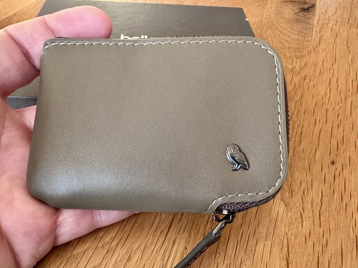 Eine Hand hält eine olivgrüne Geldbörse, ein Bellroy Card-Pocket, mit einem Reißverschluss und einem kleinen Eulen-Logo auf der Vorderseite. Im Hintergrund ist eine schwarze Box sichtbar.