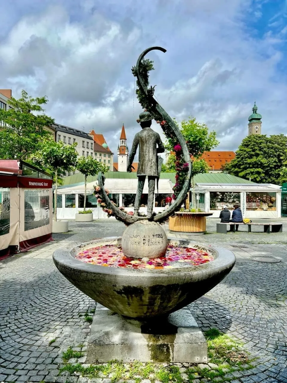 Karl Valentin Brunnen mit Blick auf den Marienplatz am Viktualienmarkt in München. Im Brunnen sind bunte Blüten von Blumen.