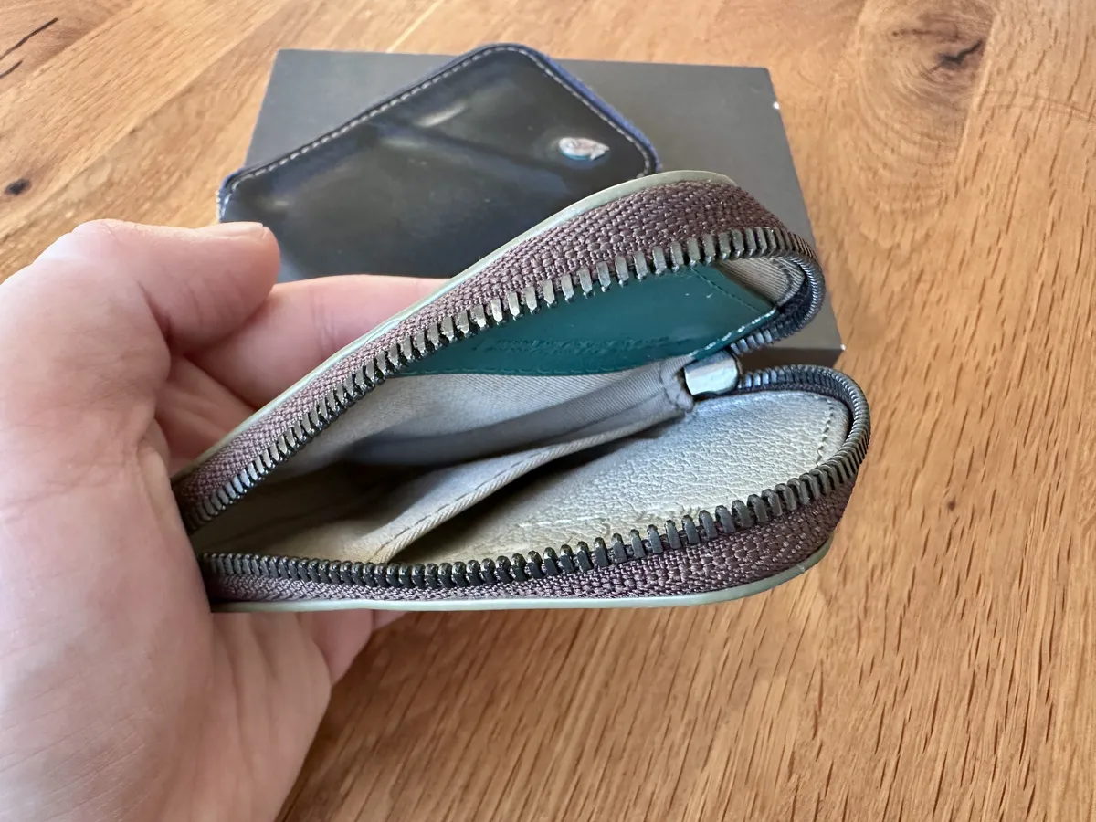 Eine Hand hält eine geöffnete Geldbörse, ein Bellroy Card-Pocket, mit einem braunen Reißverschluss. Im Hintergrund ist eine schwarze Box sichtbar.