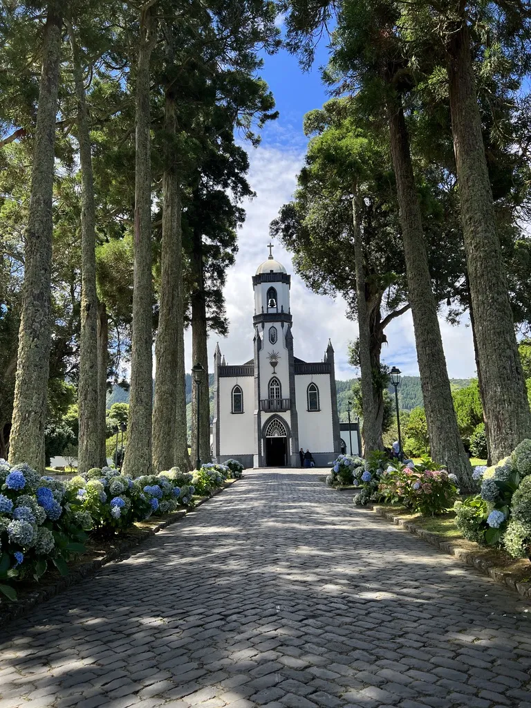 Eine elegante Kirche mit einer schwarzen und weißen Fassade auf der Azoren-Insel Sao Miguel ist am Ende eines gepflasterten Weges zu sehen, der von hohen Bäumen und blühenden Hortensien gesäumt ist. Der Himmel zeigt einige Wolken und das Gesamtbild strahlt eine ruhige, einladende Atmosphäre aus.