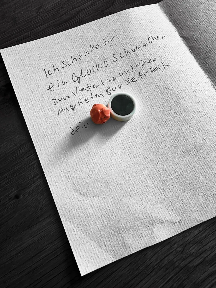 Handgeschriebene Vatertagskarte auf strukturiertem Papier mit einem kleinen rosa Glücksschweinchen und einem runden Magneten darauf. Die Nachricht lautet: „Ich schenke dir ein Glücksschweinchen zum Vatertag und einen Magneten für die Arbeit. Dein […]“
Die Szene ist liebevoll und persönlich inszeniert, aufgenommen auf einem dunklen Holztisch.