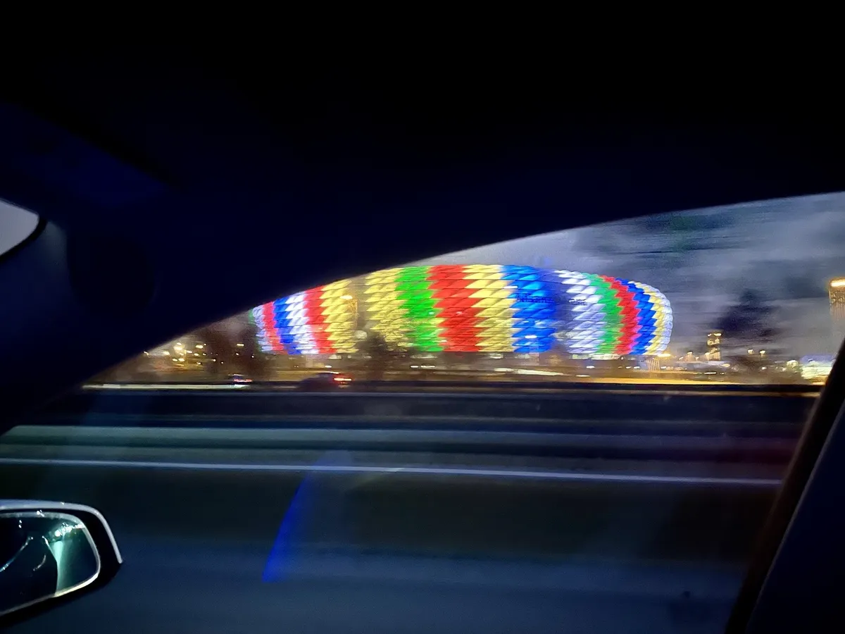 Die Allianz-Arena leuchtet nachts in bunten Farben und ist von innen durch das Fenster eines vorbeifahrenden Autos zu sehen.