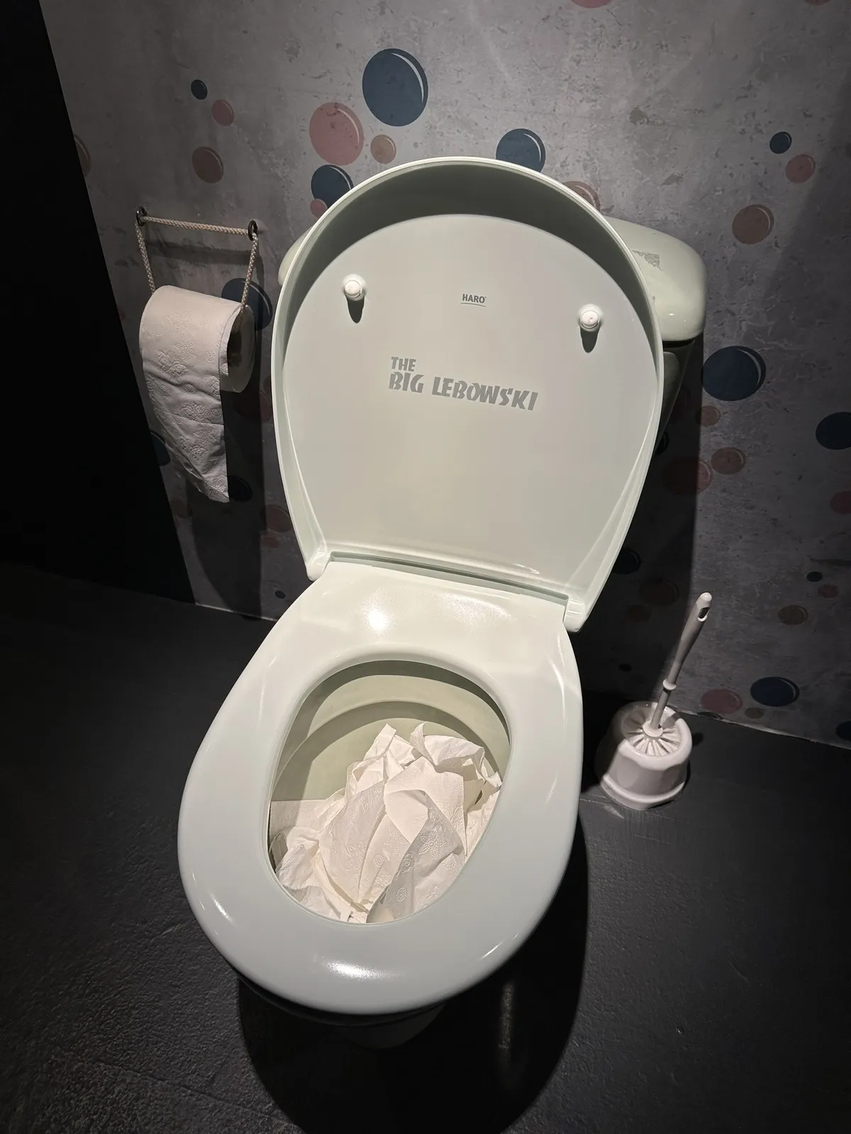 Eine hellgrüne Toilettenschüssel in einer dunklen Ausstellungsumgebung. Auf der Innenseite des hochgeklappten Deckels steht in großen Buchstaben "THE BIG LEBOWSKI". In der Schüssel liegt zusammengeknülltes weißes Papier. Daneben steht eine Toilettenbürste und an der Wand hängt eine Rolle Toilettenpapier.