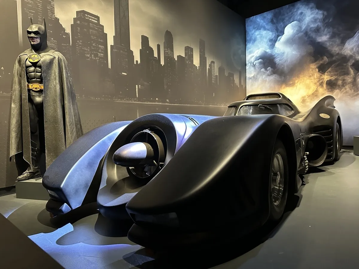 Ein Ausstellungsstück mit einem originalgroßen Batmobil in Schwarz, beleuchtet mit blauen Akzenten, vor einer beleuchteten Kulisse der Skyline von Gotham City. Links daneben steht eine Schaufensterpuppe in einem Batman-Kostüm, das dem von Michael Keaton in Batman (1989) ähnelt.
