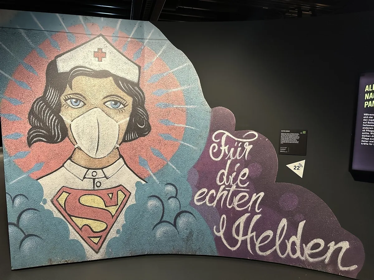 Ein Wandgemälde im Pop-Art-Stil, das eine Krankenschwester mit Mundschutz und einer weißen Haube mit rotem Kreuz zeigt. Sie trägt ein rotes Superman-Logo auf der Brust. Rechts neben der Figur steht in geschwungener, weißer Schrift auf lilafarbenem Untergrund der Satz "Für die echten Helden".