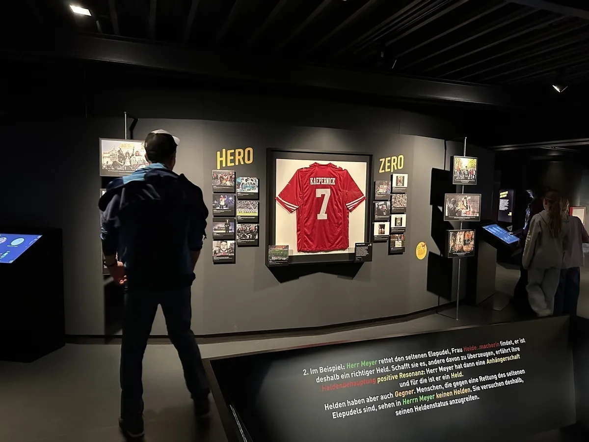 Ein dunkler Ausstellungsraum, in dem Besucher vor einer Wand mit der Aufschrift "HERO | ZERO" stehen. Im Zentrum ist ein gerahmtes, rotes Fußballtrikot mit der Nummer 7 und dem Namen "KÄFERSTEIN" ausgestellt, umgeben von kleinen Bildern und Videoscreens. Am Boden ist ein Infotext in deutscher Sprache zu sehen.