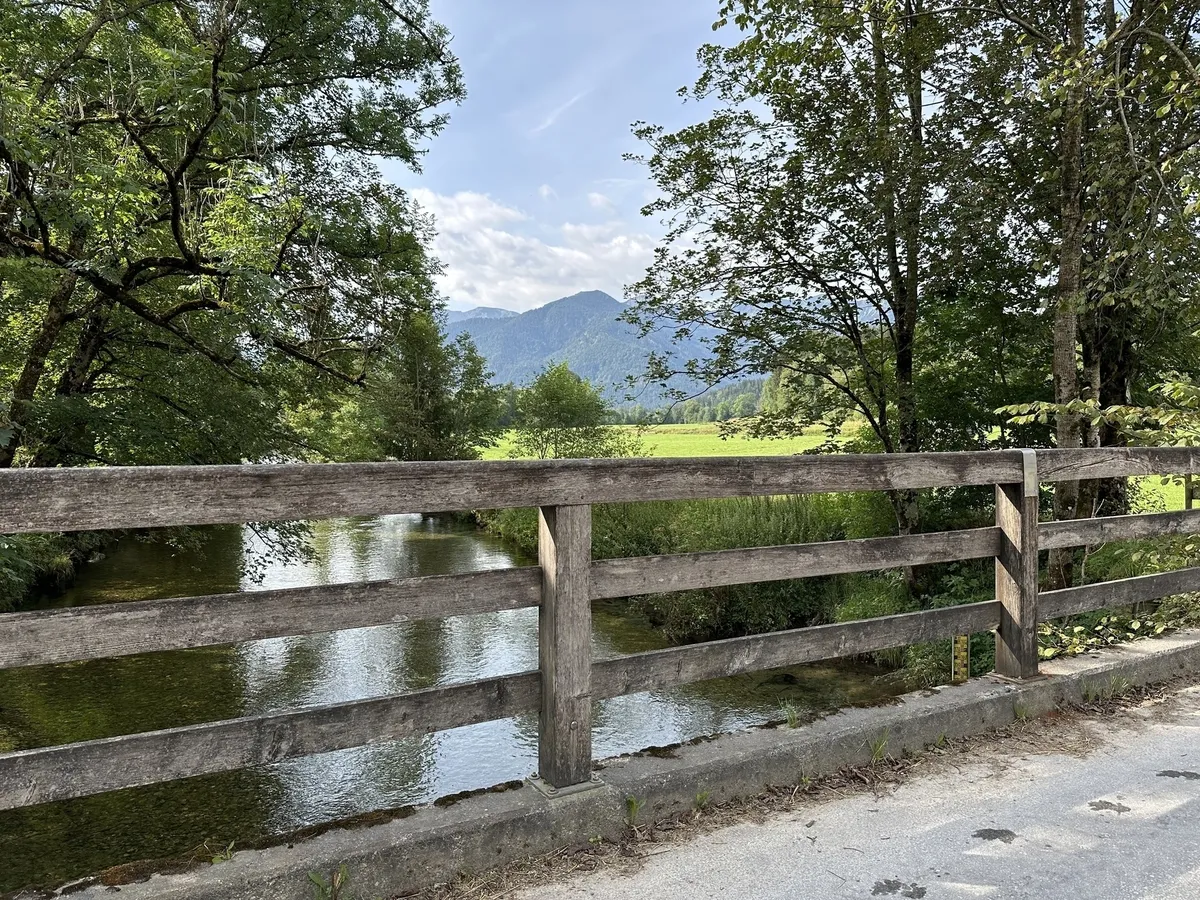 Eine Holzbrücke über einen kleinen, klaren Fluss, der durch eine von Bäumen gesäumte Wiese fließt. Im Hintergrund sind grüne Berge unter einem leicht bewölkten Himmel zu sehen.