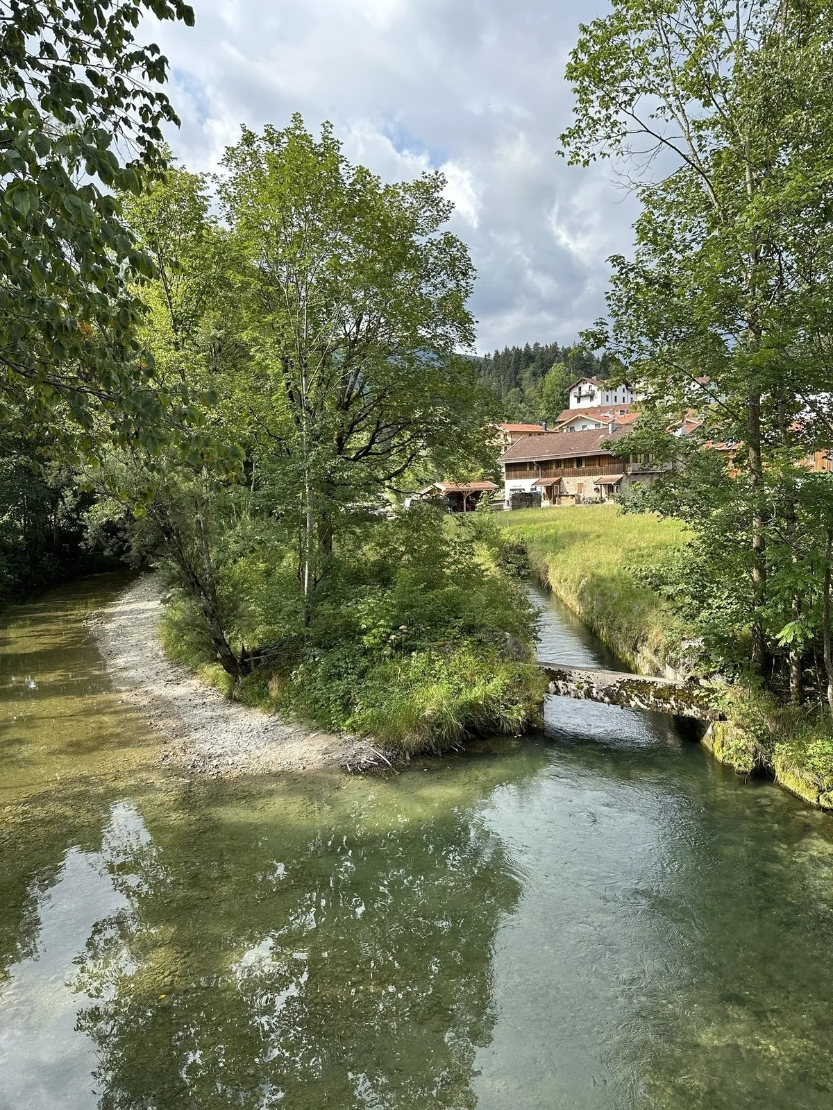 Ein kleiner Fluss mit einer bewachsenen Insel in der Mitte, über die eine kleine, rustikale Steinbrücke führt. Am Ufer stehen große Bäume. Im Hintergrund sind traditionelle Häuser mit braunen Holzbalkonen und roten Dächern auf einer Anhöhe zu sehen.