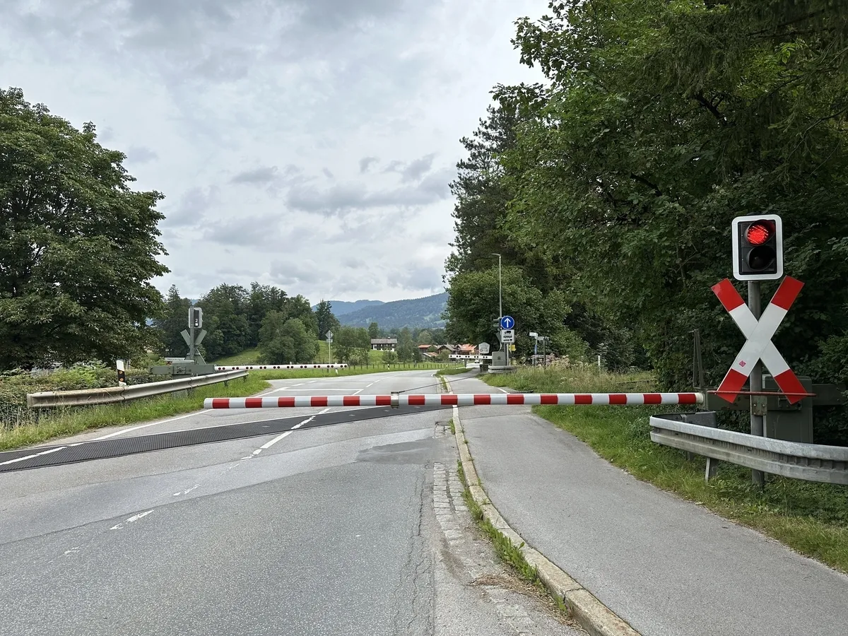 Ein geschlossener Bahnübergang mit einer rot-weißen Schranke und einer rot leuchtenden Warnleuchte sowie einem Andreaskreuz. Die Straße führt in eine baumbestandene Landschaft mit Häusern im Hintergrund.
