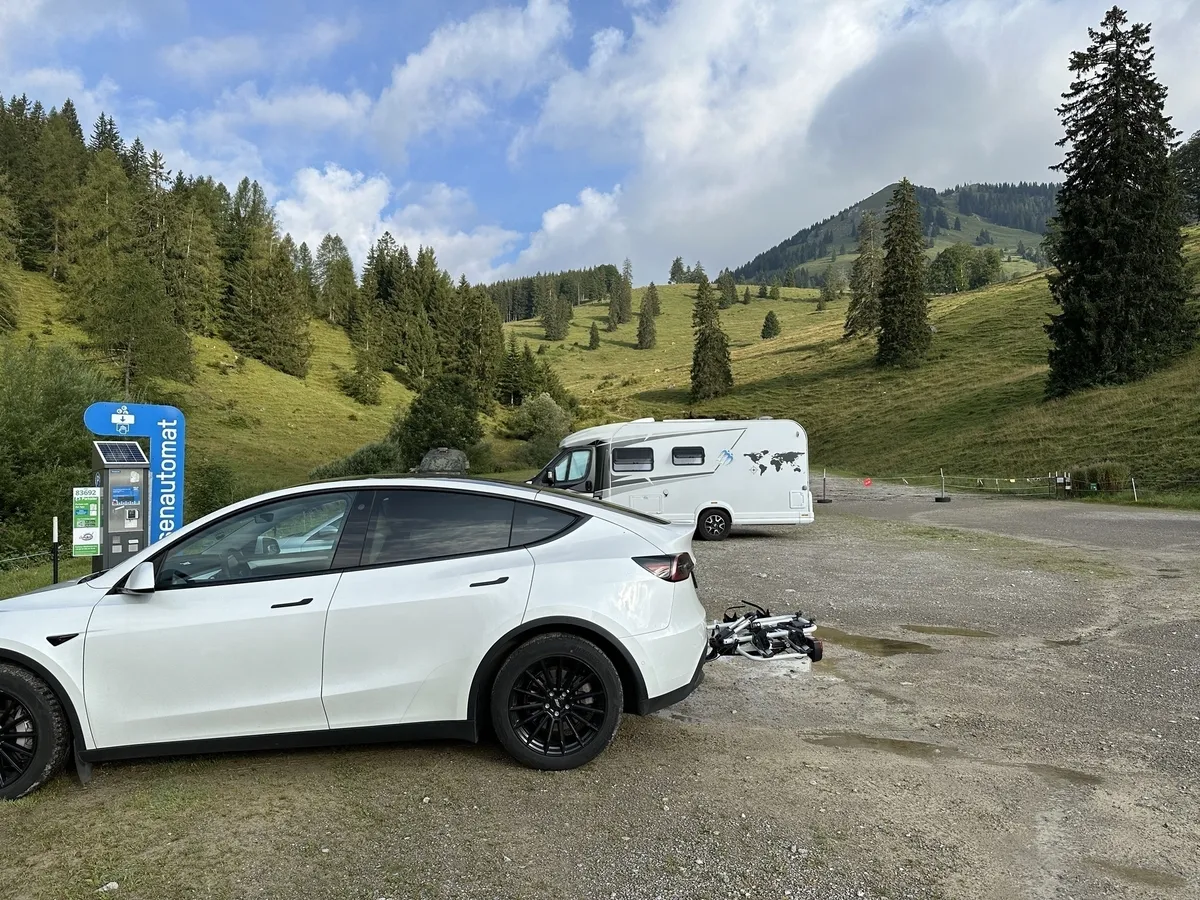 Ein weißes Tesla Model Y mit Anhängerkupplung und ein weißes Wohnmobil auf einem Schotterparkplatz vor einer grünen, bergigen Landschaft mit Bäumen und Wiesen. Links steht ein blauer Parkscheinautomat.