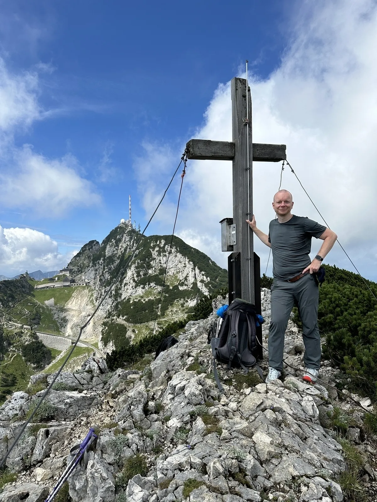 René Fisher in dunkler Wanderkleidung steht auf einem felsigen Berggipfel neben einem Gipfelkreuz. Im Hintergrund erstreckt sich eine felsige Bergkette mit dem Wendelstein, auf deren höchstem Gipfel eine große Funk-/Sendeanlage zu erkennen ist, unter einem blauen Himmel mit weißen Wolken.