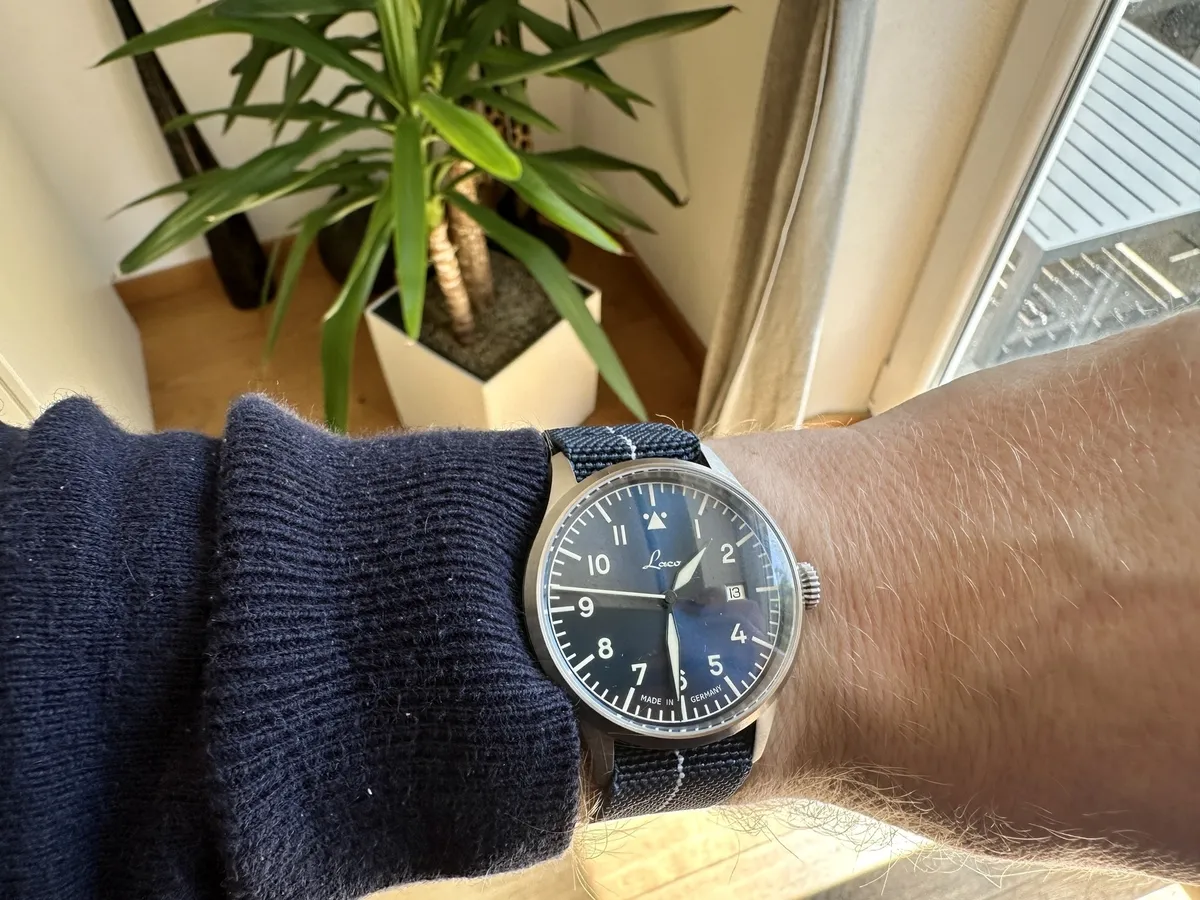 Ein Mann trägt eine Laco Stuttgart Pro mit einem blauen Zifferblatt und einem dunkelblauen Armband, während im Hintergrund eine Zimmerpflanze steht.