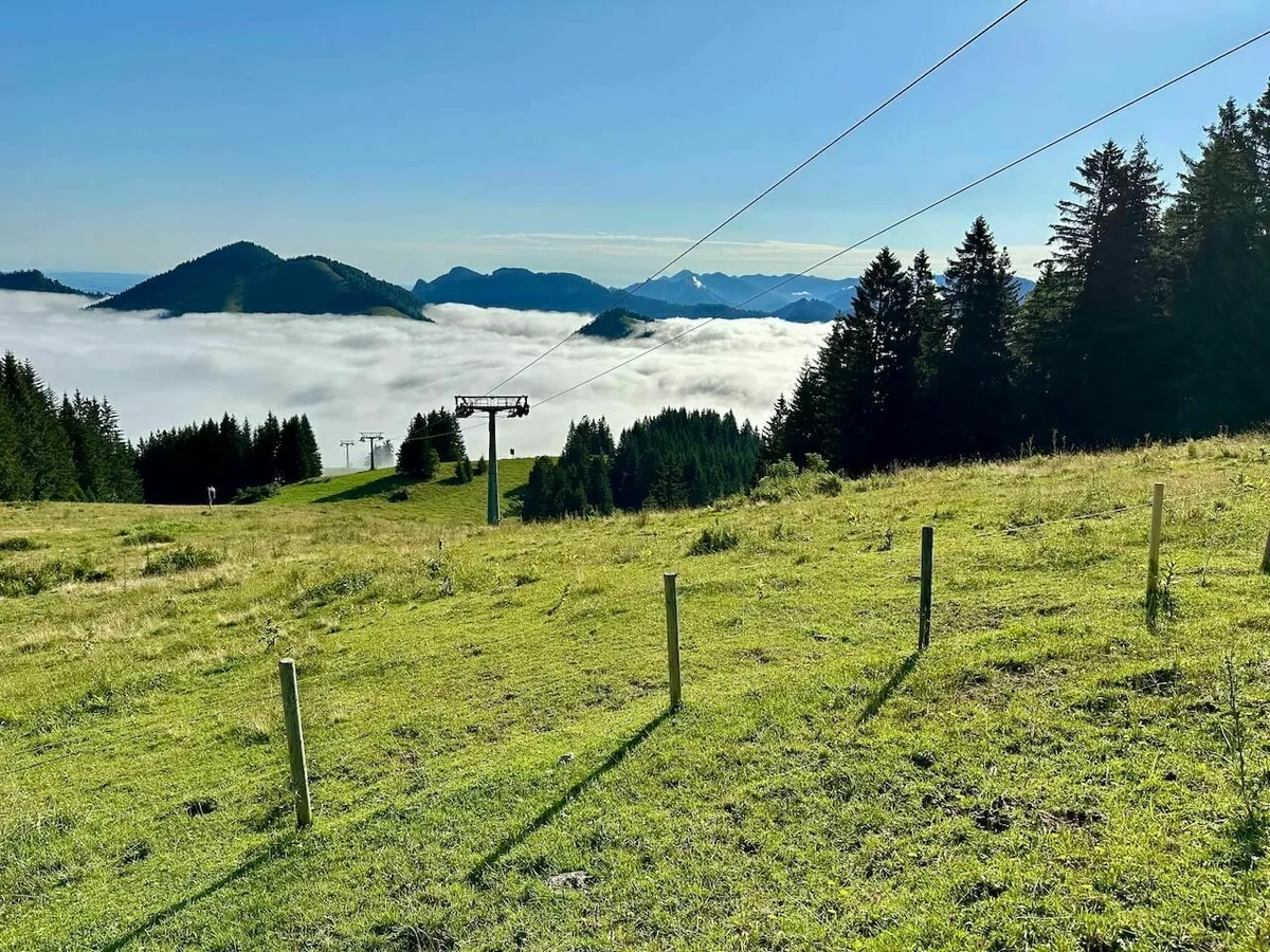 Grüne Bergwiese mit Zaunpfosten und einer Seilbahnstütze, die über ein dichtes Wolkenmeer (Nebelmeer) im Tal ragt, mit Bergspitzen am Horizont