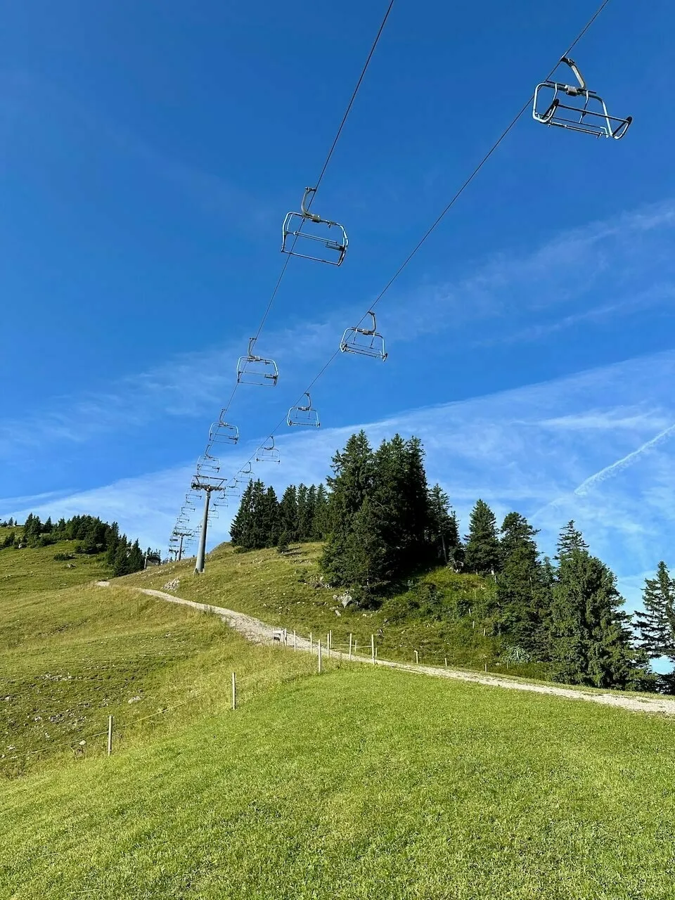 Blick bergauf auf eine Doppelsesselbahn über einer grünen Skiwiese mit einem Schotterweg, vor klarem blauem Himmel