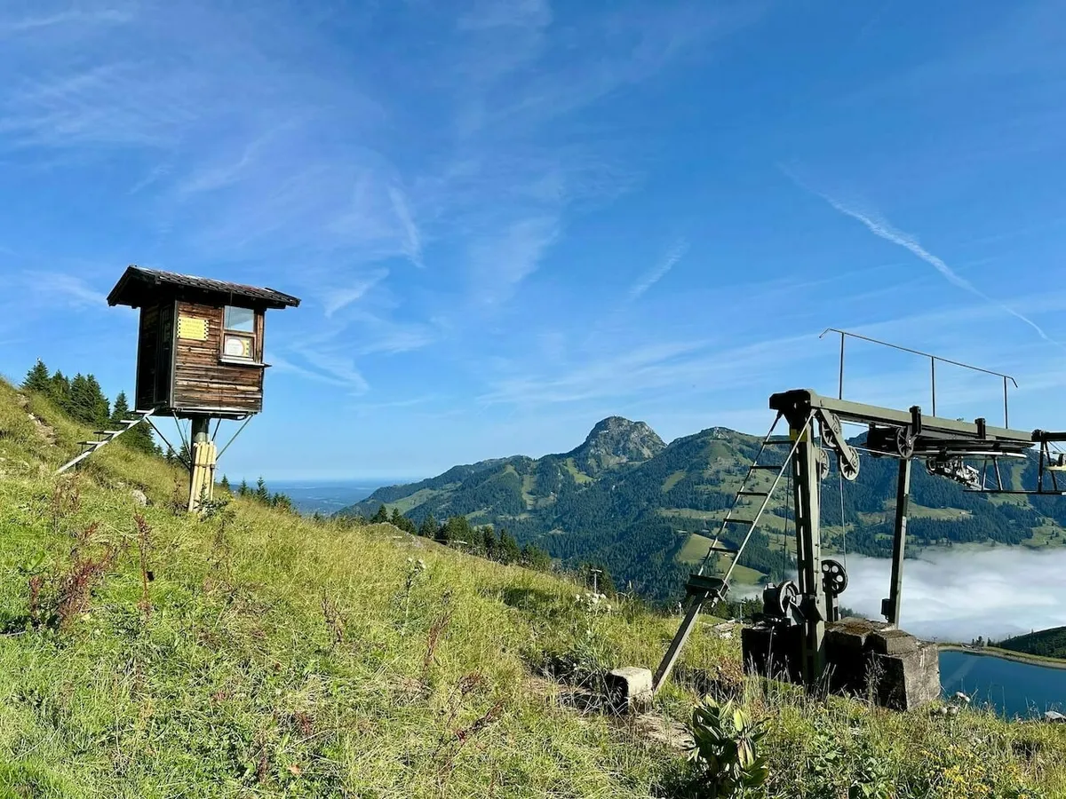 Kleine hölzerne Hütte auf Stelzen neben einer alten, metallenen Liftanlage auf dem Berg, mit Blick über das Tal und einen Stausee