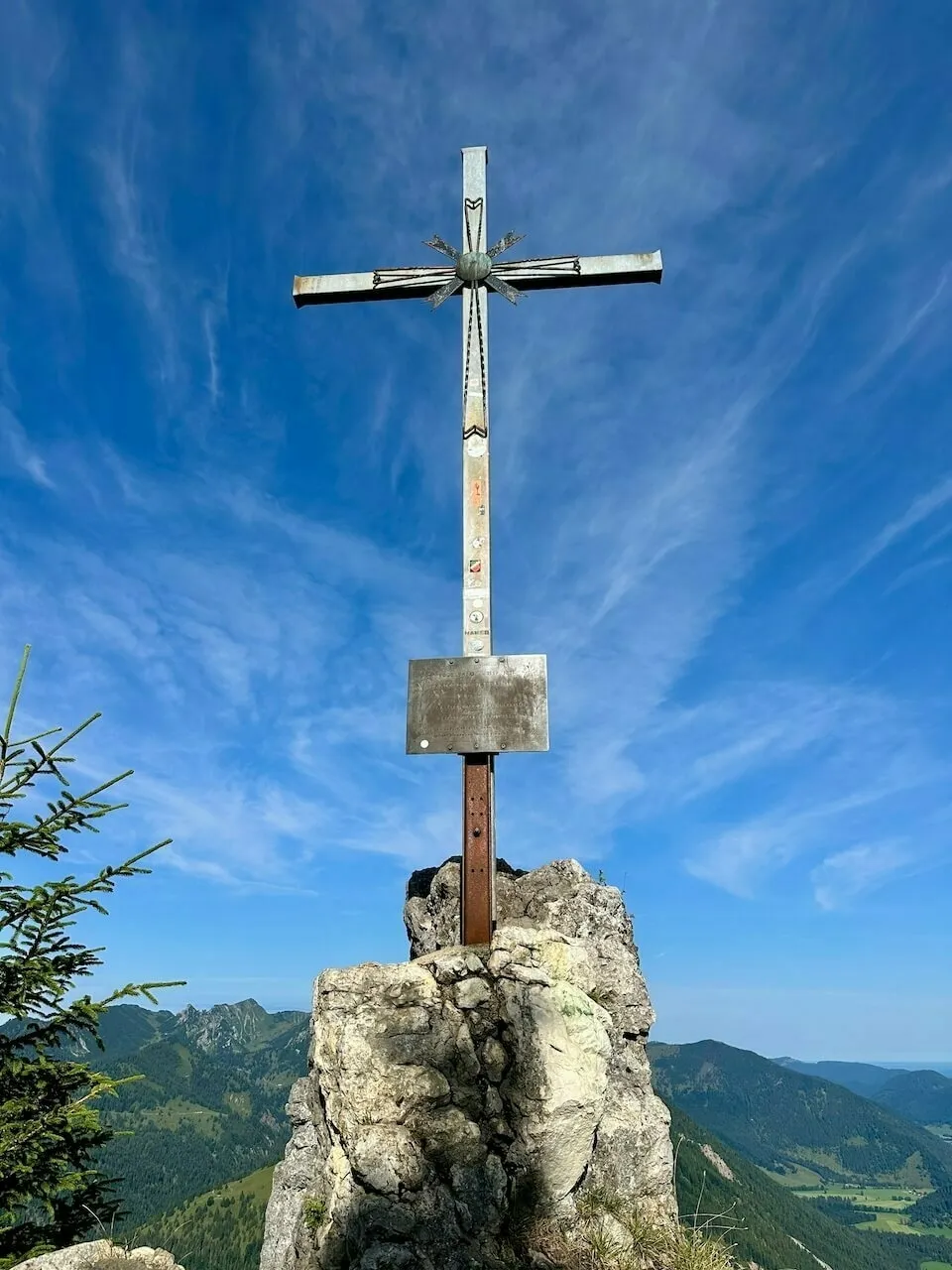Monumentales Metall-Gipfelkreuz mit Verzierungen auf einem Felsen, im Hintergrund blauer Himmel mit Schleierwolken und grüne Bergketten
