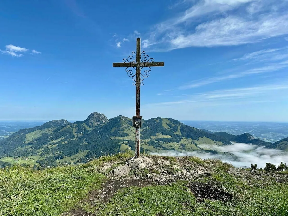 Historisches, schmiedeeisernes Gipfelkreuz auf einem grasbewachsenen Felsvorsprung, mit Blick über grüne Berge, Nebelfelder und eine weite Ebene