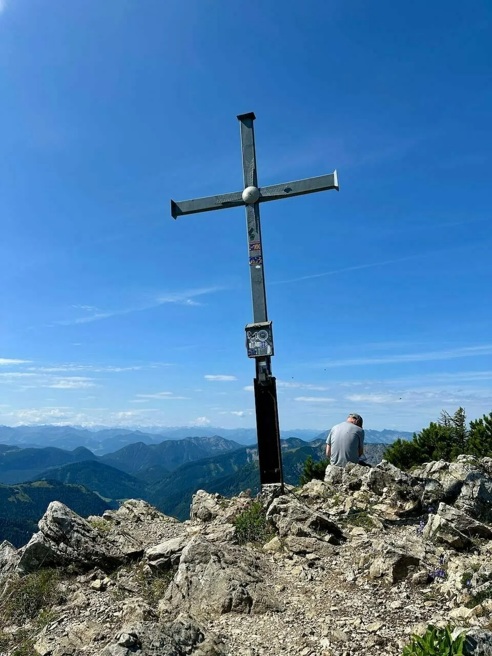 Männlicher Wanderer sitzt neben einem Metall-Gipfelkreuz auf einem felsigen, kargen Gipfel unter klarem blauem Himmel, mit Fernblick auf Bergketten