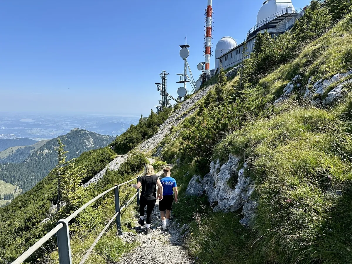 Zwei Personen wandern auf einem Bergpfad neben einer Forschungsstation mit Antennen und Kuppeln, umgeben von grüner Vegetation und einem weiten Blick auf die Landschaft.