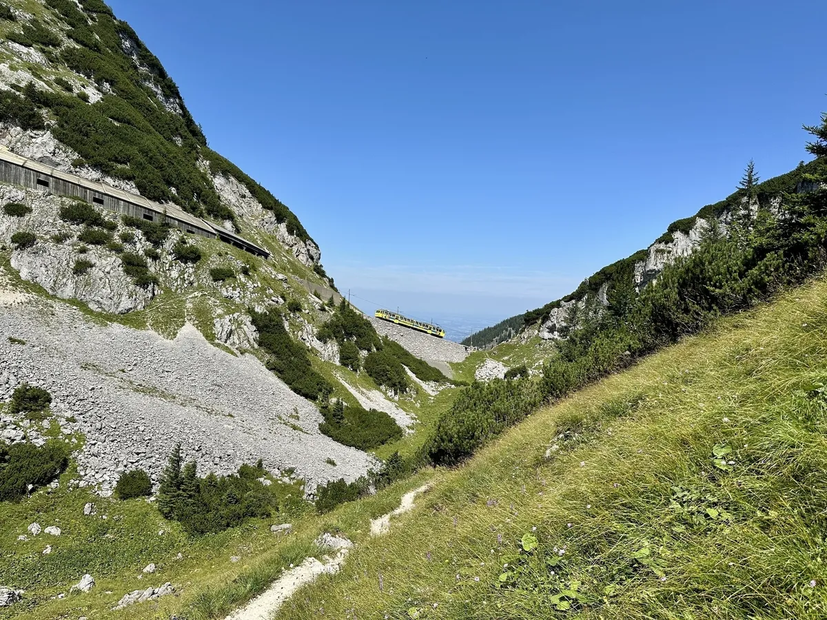 Die Wendelsteinzahnradbahn fährt entlang einer Gebirgsstrecke, die von grünen Hügeln und Felsen umgeben ist.