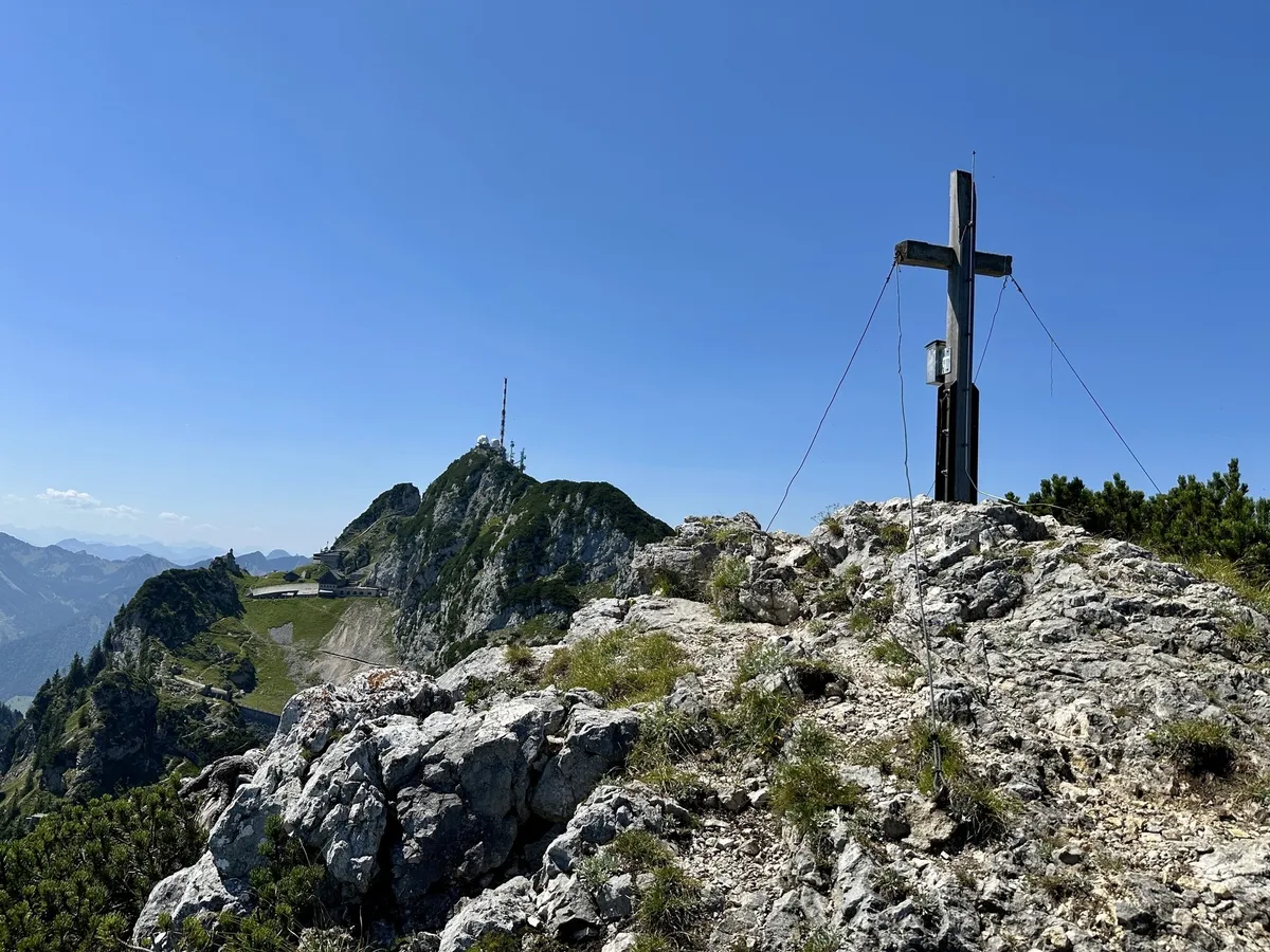 Ein Gipfelkreuz steht auf einem felsigen Berggipfel, mit einem Sender in der Ferne und klarem blauen Himmel.