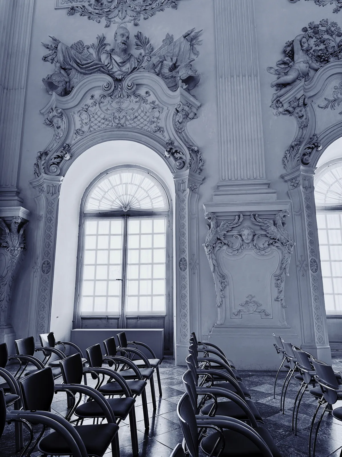 Ein prachtvoll verzierter klassischer Saal mit hohen Bögen, detaillierten Stuckarbeiten und einer Reihe moderner Stühle im Schloss Schleißheim.