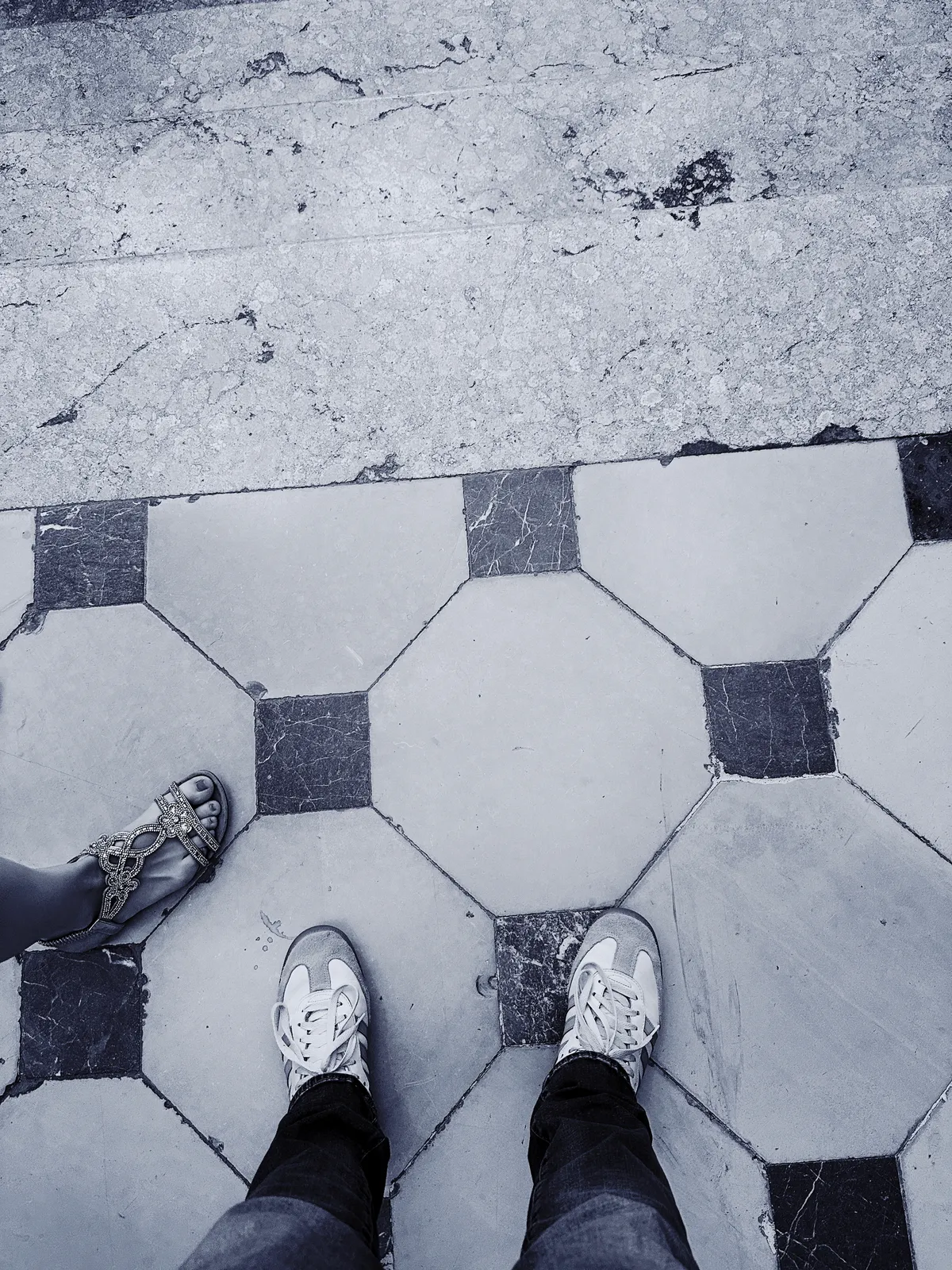 Zwei Personen stehen auf einem Boden mit einem schwarz-weißen sechseckigen Muster, wobei eine Person Sandalen und die andere Sneakers trägt.