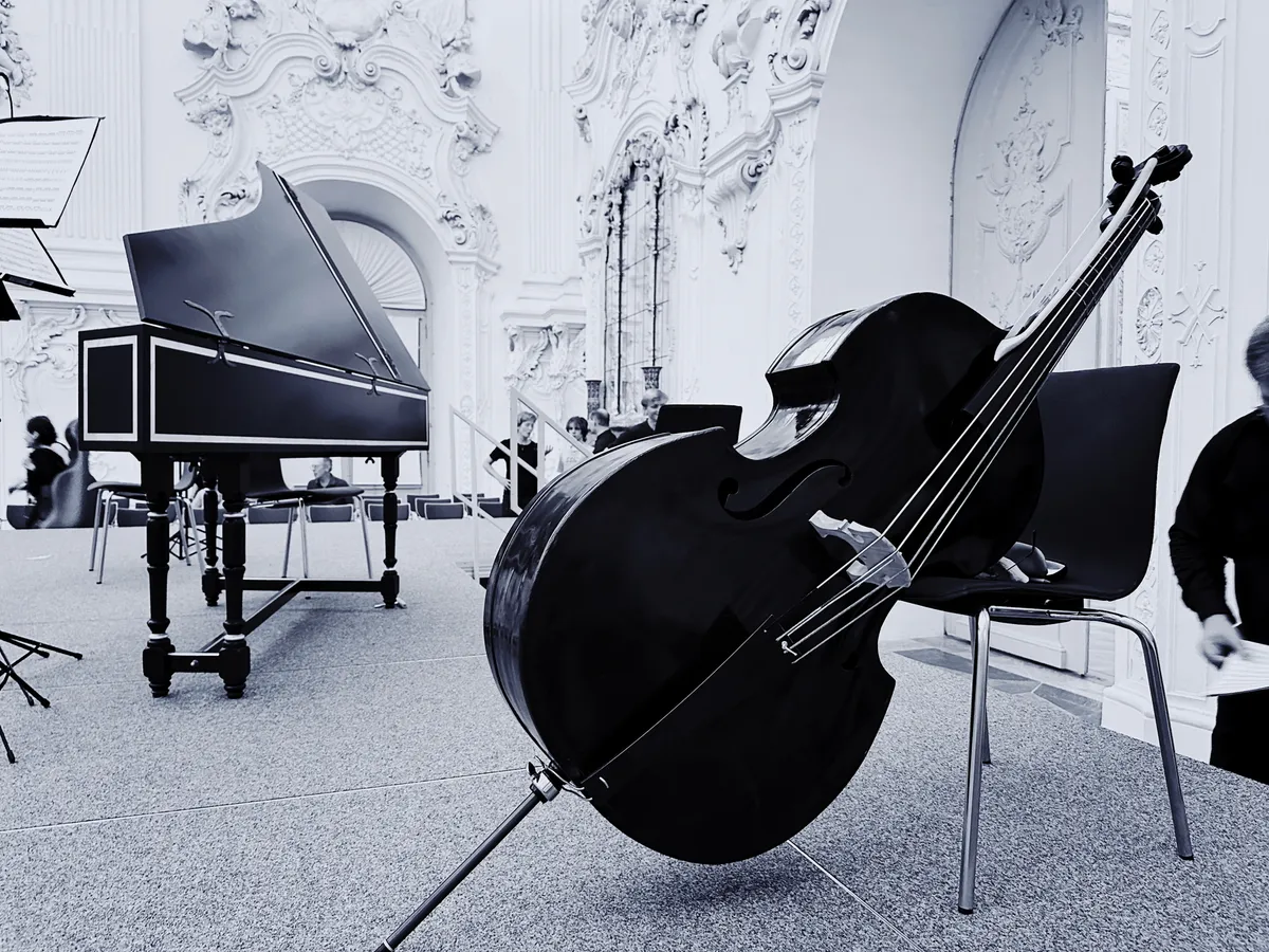 Ein Kontrabass lehnt an einem Stuhl auf einer Bühne neben einem historischen Tasteninstrument in einem reich verzierten Raum.