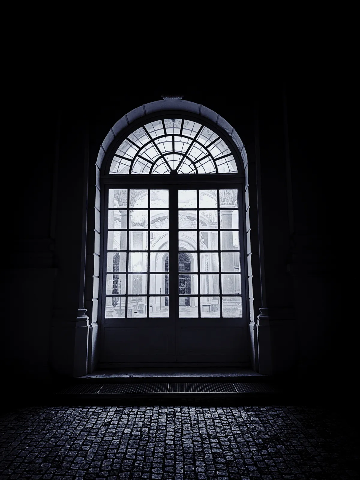 Ein großes, bogenförmiges Fenster mit Glaspaneelen lässt schwaches Licht in einen dunklen Raum auf einem Kopfsteinpflasterboden strömen.