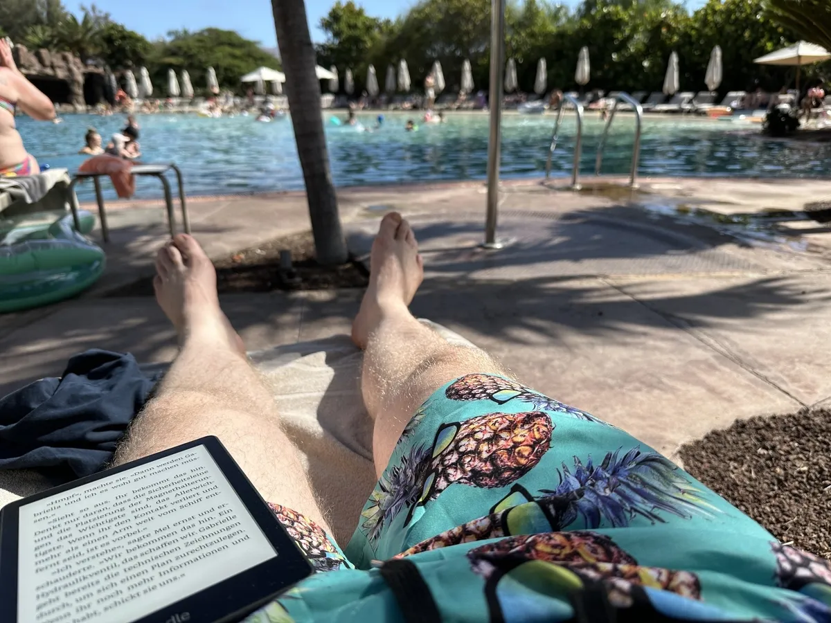 Eine Person entspannt sich auf einer Sonnenliege am Pool und liest auf einem E-Reader, während andere Menschen im Wasser schwimmen.