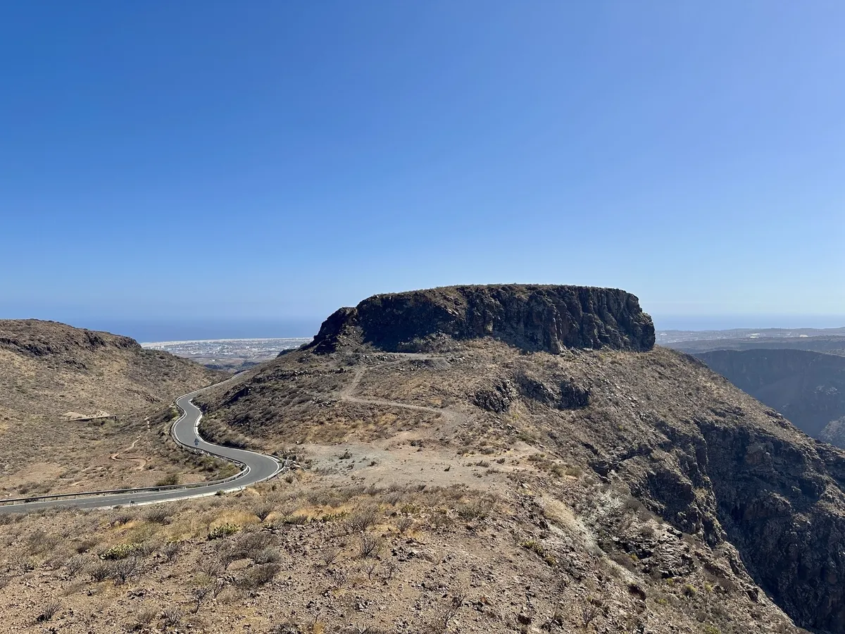Eine kurvenreiche Straße auf Gran Canaria führt durch eine trockene, felsige Landschaft mit einem markanten, flachen Hügel im Hintergrund.
