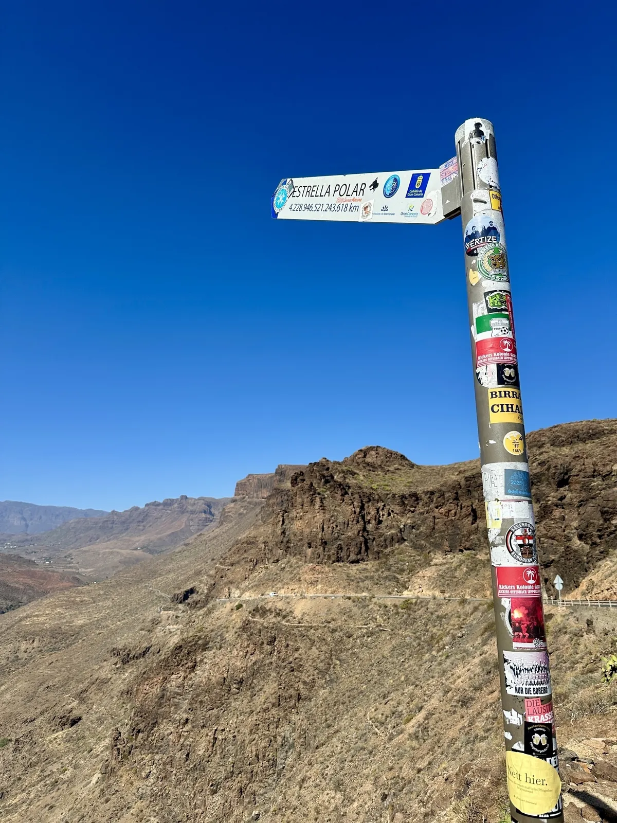 Ein mit Aufklebern bedecktes Straßenschild mit der Beschriftung ESTRELLA POLAR steht in einer bergigen, kargen Landschaft unter klarem, blauem Himmel am Miradores Astronómicos de Gran Canaria.