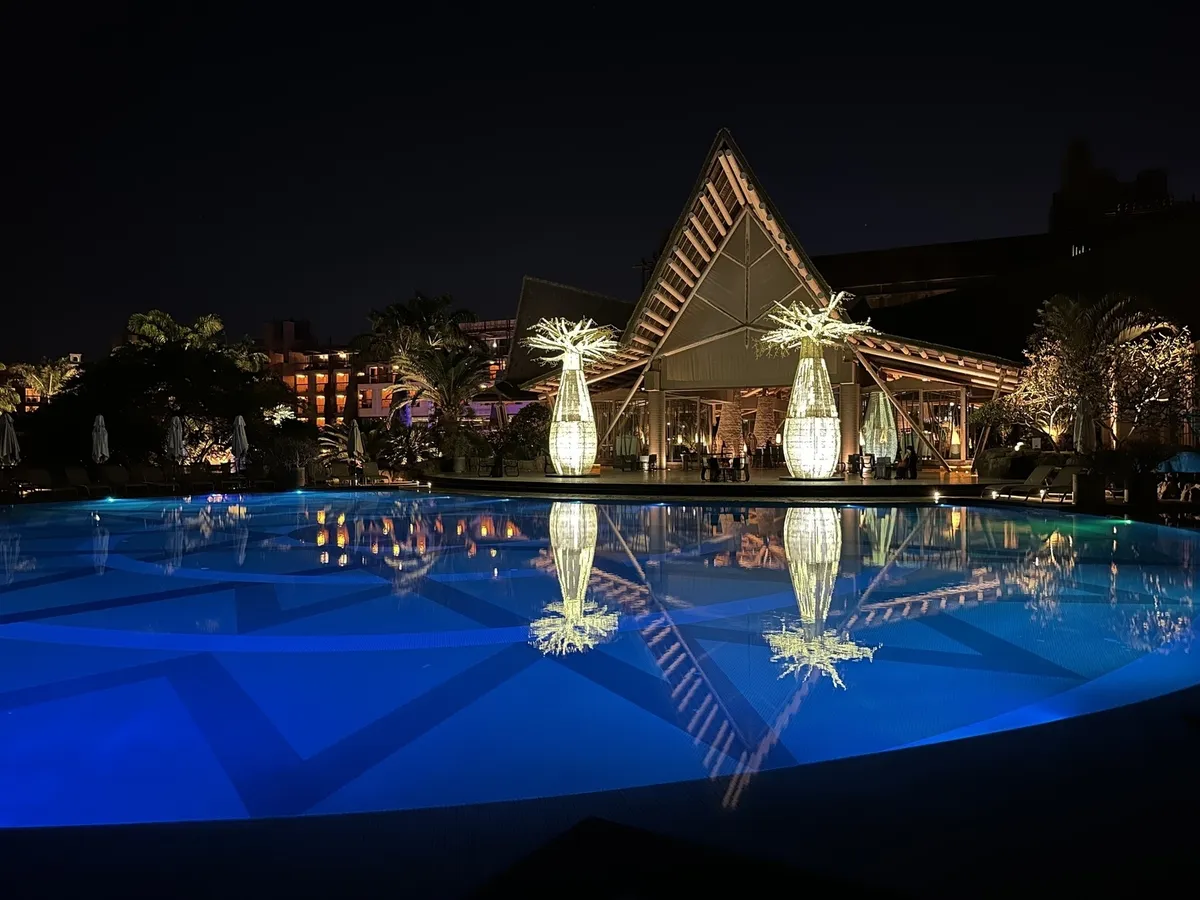 Ein beleuchteter Pool spiegelt ein modernes Gebäude und palmenartige Skulpturen in einer nächtlichen Szene wider.