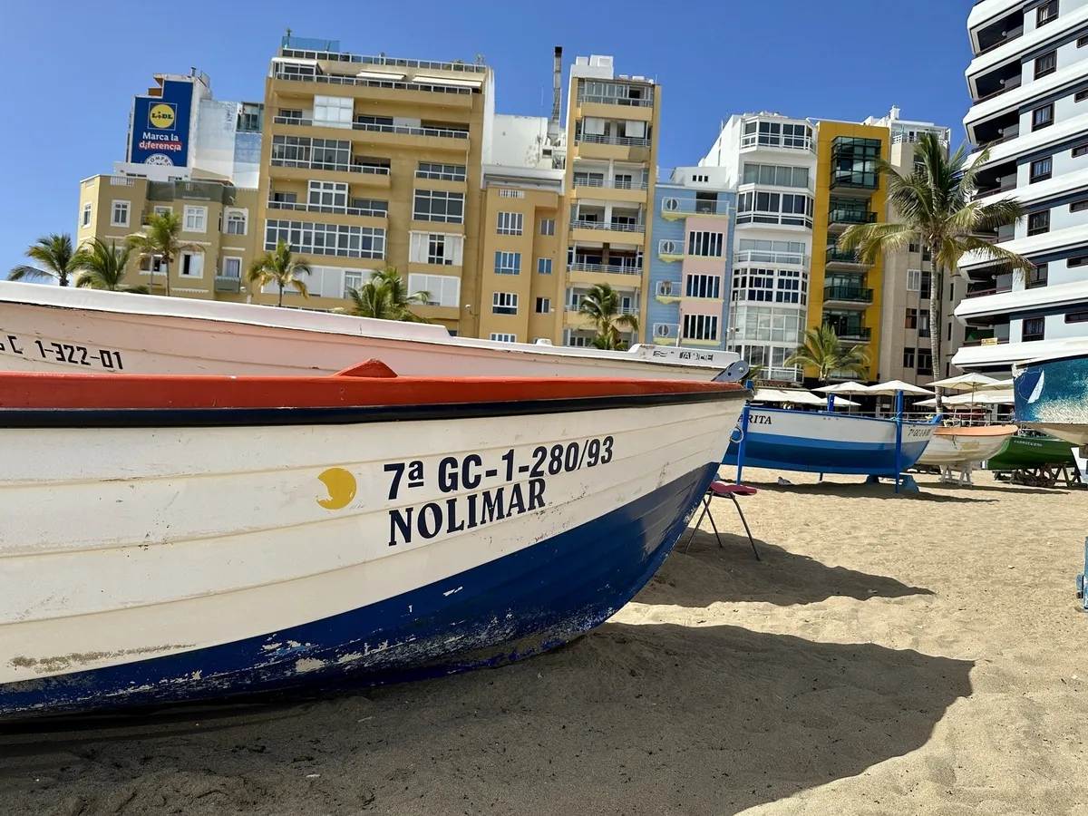 Ein Fischerboot steht am Sandstrand vor einer Reihe bunter Gebäude und Palmen.