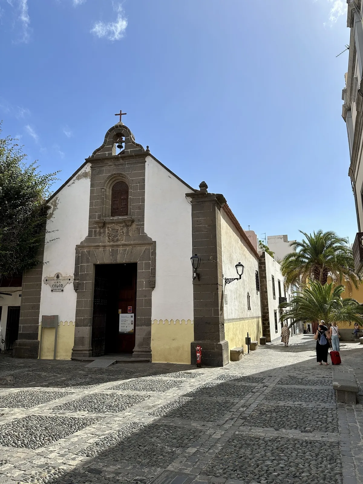 Eine historische Kirche mit schlichter Architektur steht in einer gepflasterten Straße, umgeben von Palmen und einem klaren blauen Himmel.