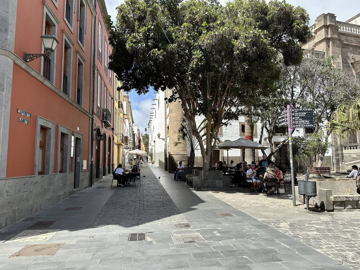 Eine malerische Straße mit bunten Gebäuden, wo Menschen in einem Café unter Bäumen sitzen.