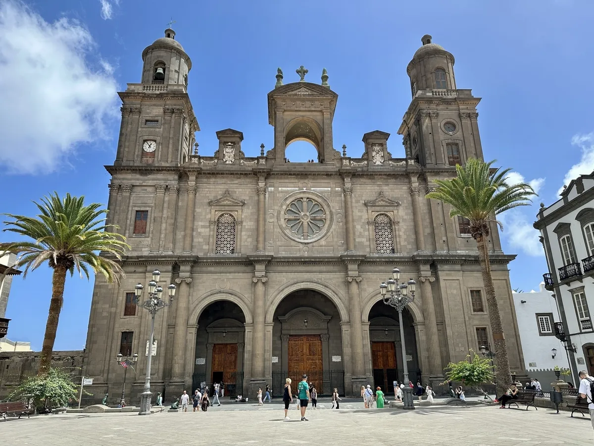 Eine beeindruckende Kathedrale mit zwei Türmen und einer großen Fassade steht auf einem weitläufigen Platz, umgeben von Palmen und besuchenden Menschen.