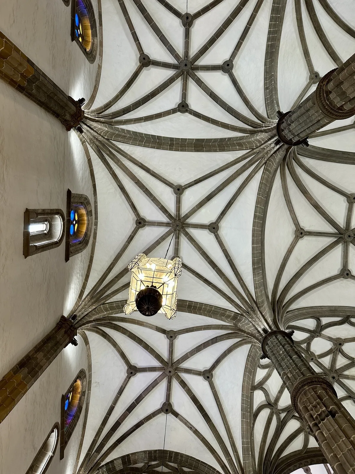 Ein kunstvoll gestaltetes gotisches Gewölbe mit farbigen Glasfenstern und einer hängenden Lampe ist zu sehen.