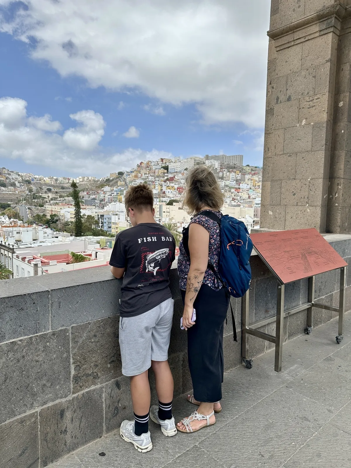 Eine Frau und ein Junge schauen von einer erhöhten Plattform auf eine malerische Stadt mit hügeligen Häusern im Hintergrund.