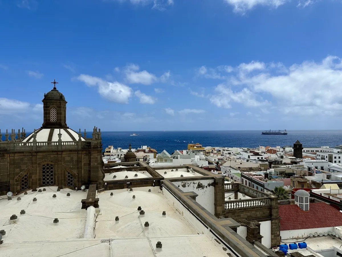 Stadtlandschaft mit Gebäuden, einem historischen Turm und dem Meer im Hintergrund unter blauem Himmel.