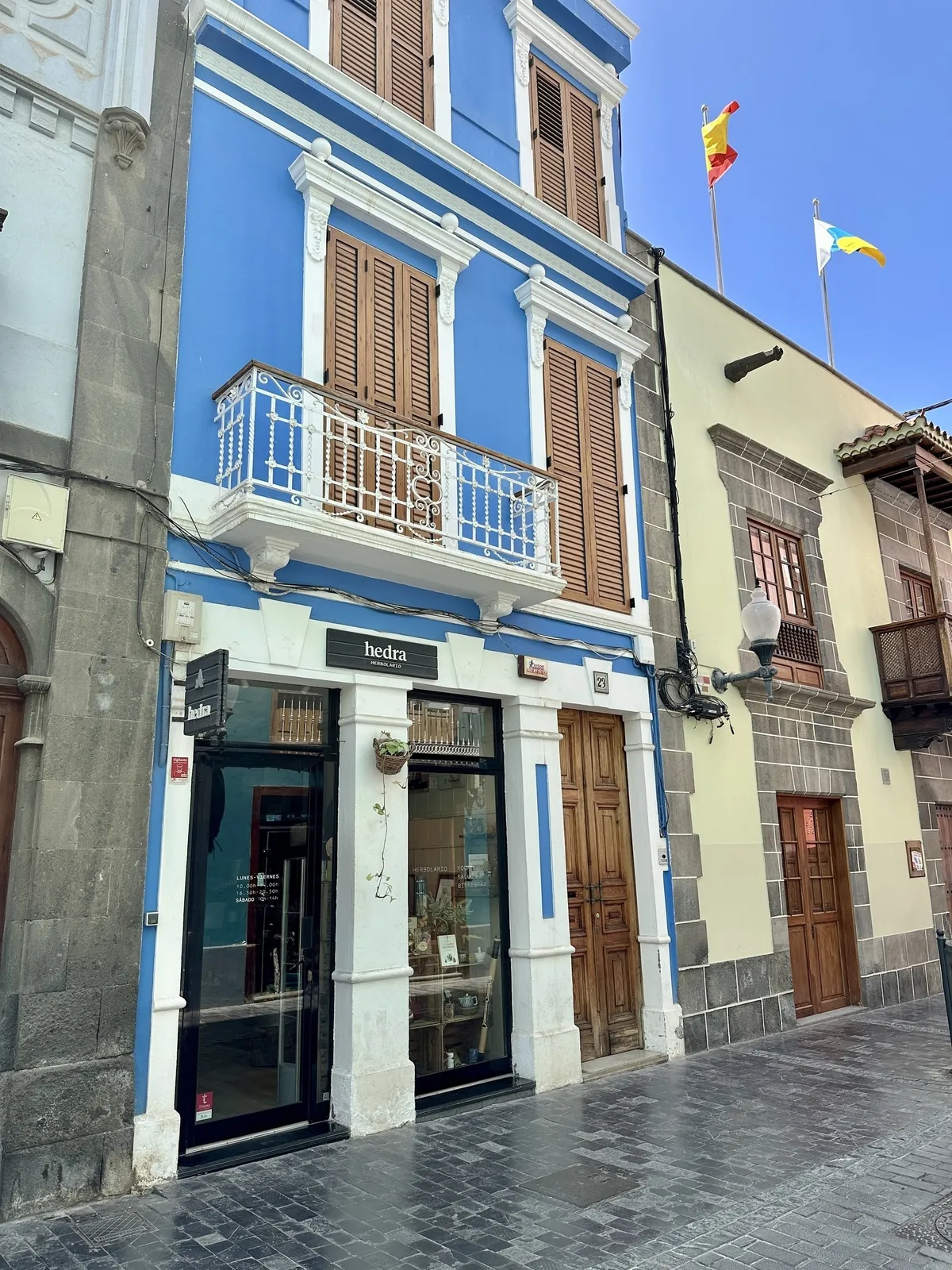 Ein blau gestrichenes Gebäude mit Holzbalkonen und zwei Etagenfenstern steht neben einem steinernen Gebäude, mit spanischen Fahnen auf dem Dach.