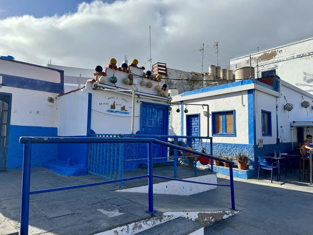Ein kleines, blau-weiß gestrichenes Gebäude mit bunten Bojen auf dem Dach steht auf einem sonnigen Platz.