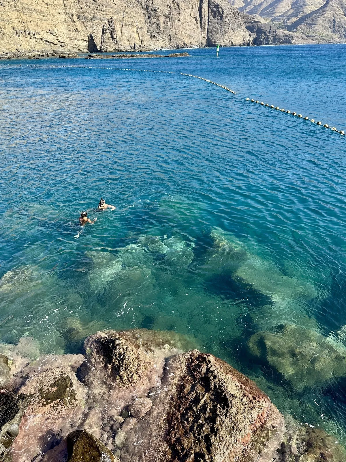 Zwei Personen schwimmen in klarem, blauem Wasser nahe einer felsigen Küste mit steilen Klippen im Hintergrund.