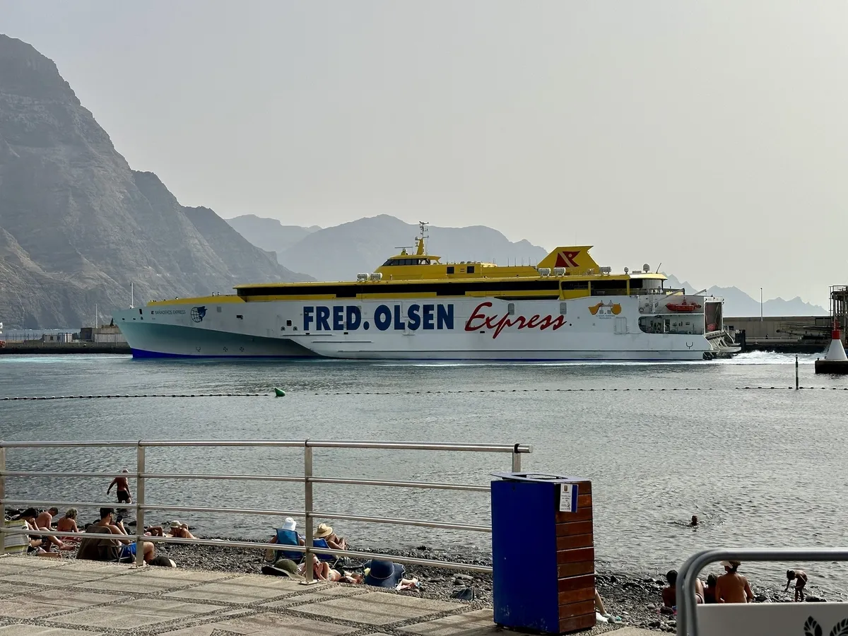 Ein großes Passagierfähre mit der Aufschrift FRED. OLSEN Express fährt in der Nähe eines Strandes mit im Wasser spielenden Menschen, während im Hintergrund hohe Berge zu sehen sind.
