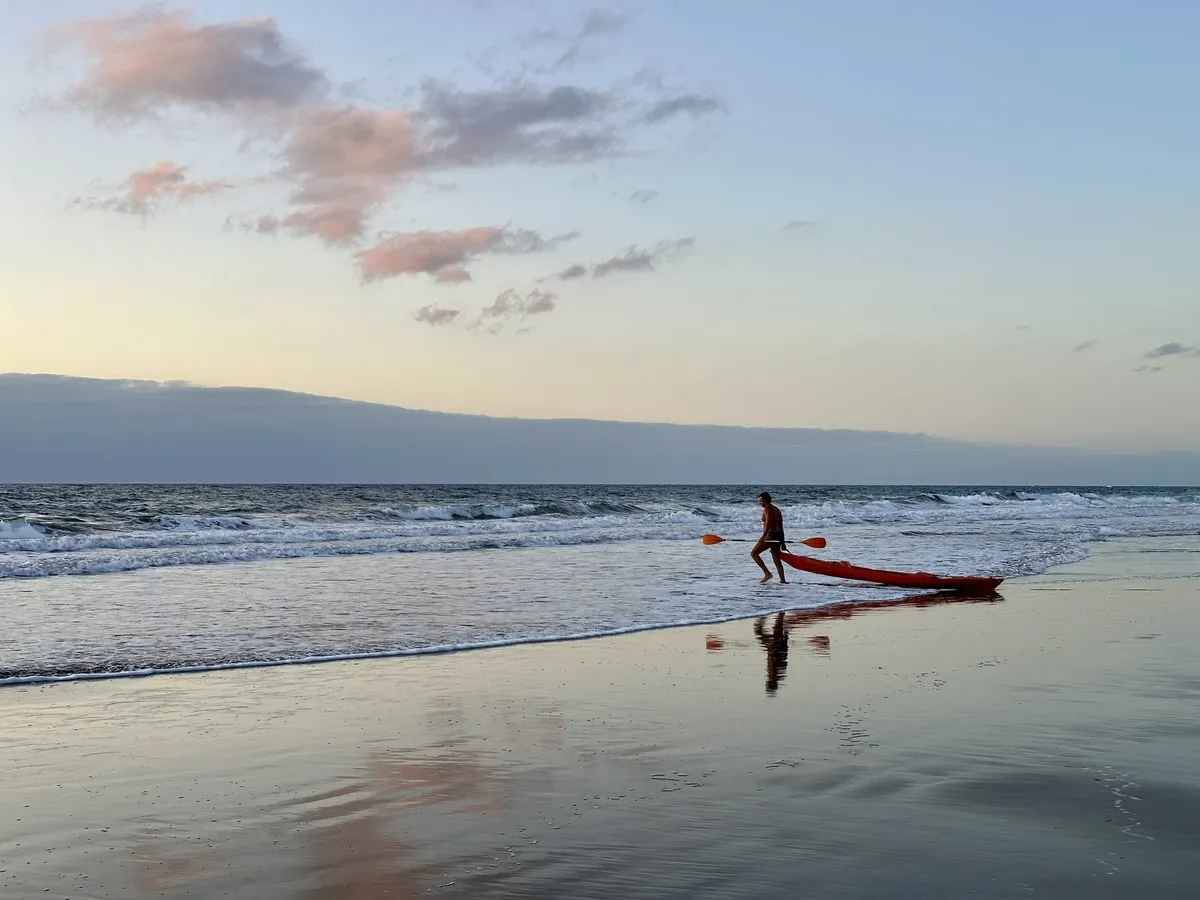 Eine Person zieht bei Sonnenuntergang ein rotes Kajak am Strand entlang des ruhigen Meeres.