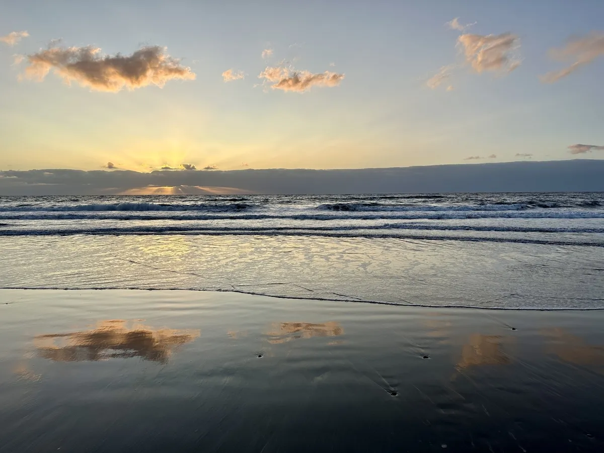 Ein Sandstrand bei Sonnenuntergang mit Wolken am Horizont und Spiegelungen im Wasser.