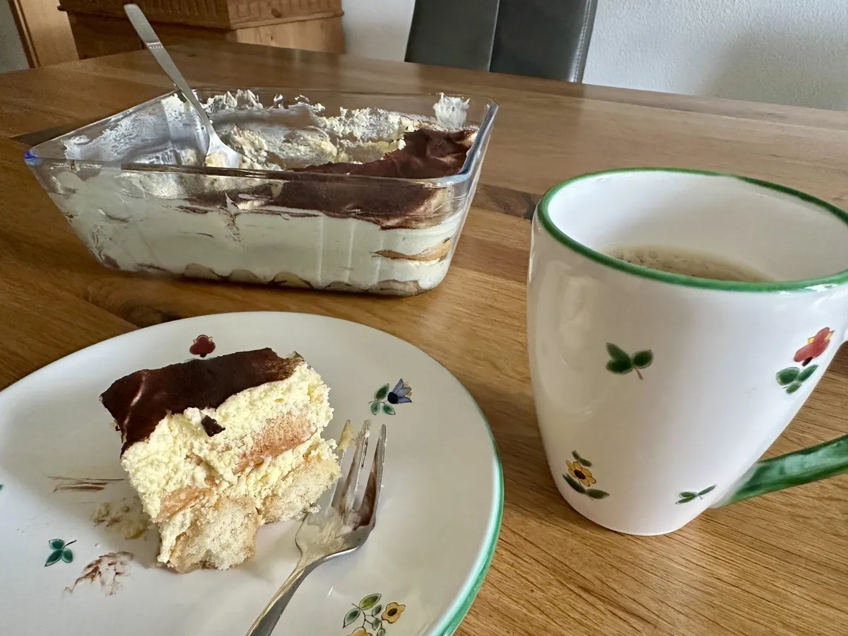 Ein Stück Tiramisu auf einem Teller neben einer großen Tasse Kaffee und einer Schale mit mehr Tiramisu stehen auf einem Holztisch.