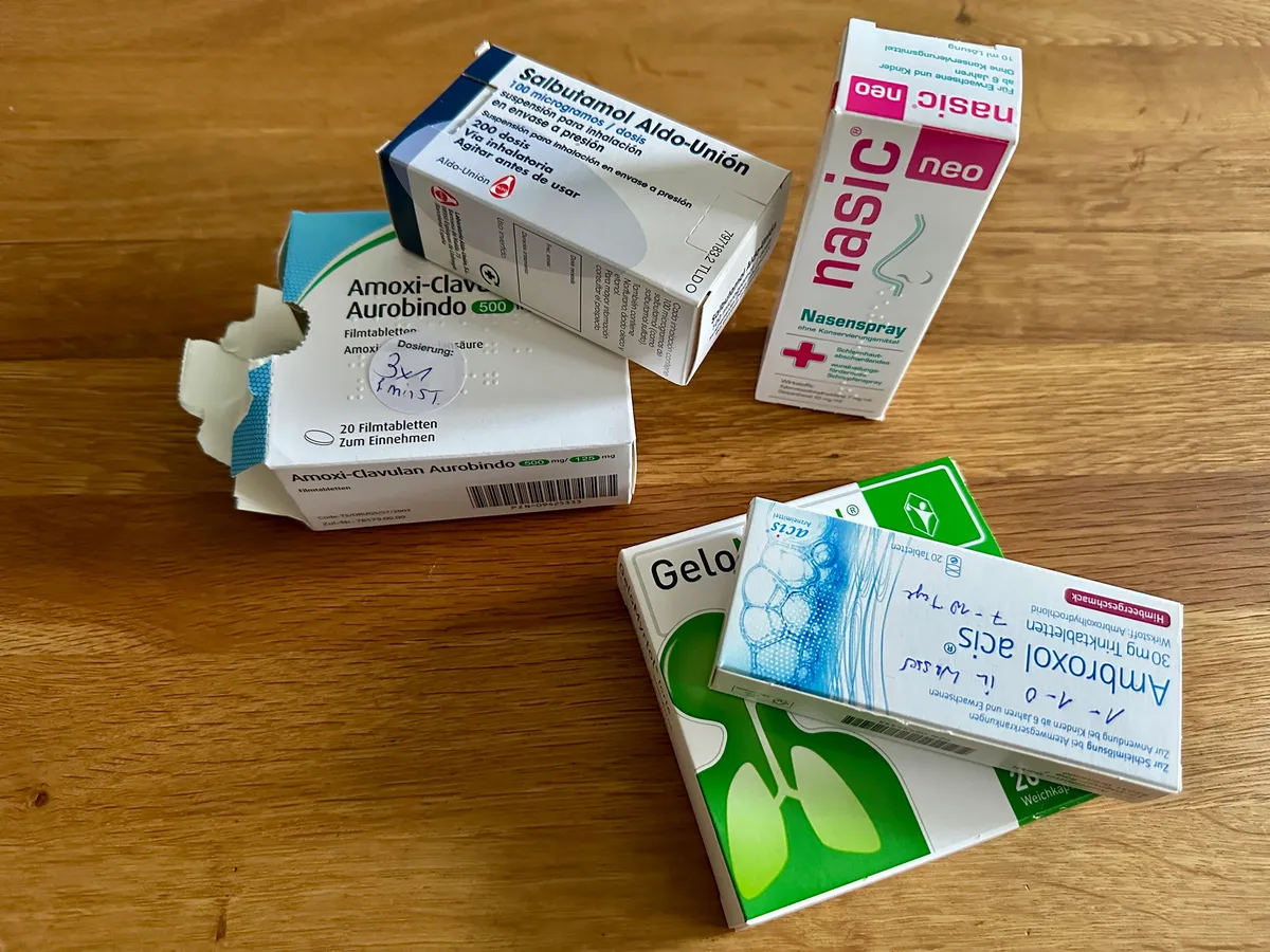 Auf einem Holztisch liegen verschiedene Medikamentenschachteln, darunter Antibiotika und Nasenspray.