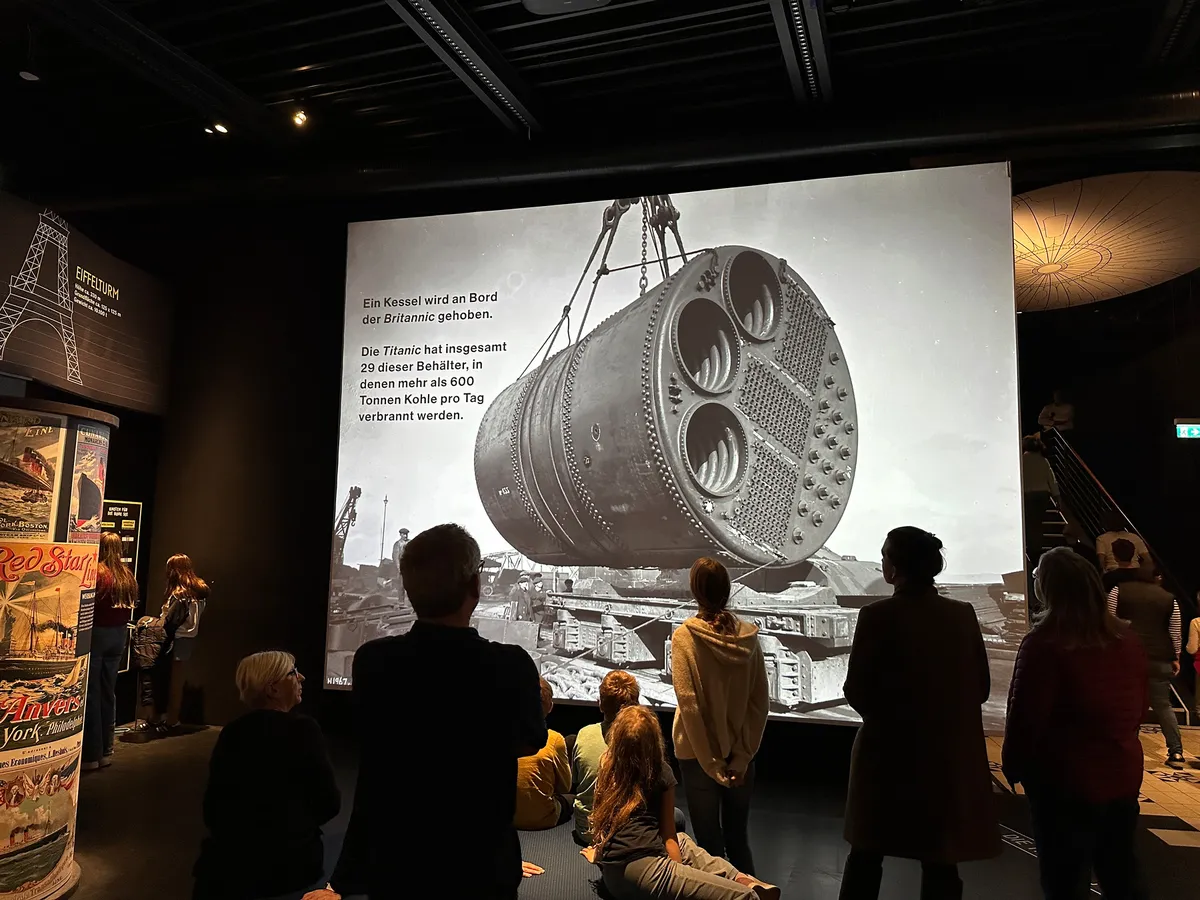 Eine Gruppe von Menschen betrachtet ein großes historisches Foto von einem Kessel der Titanic, das in einem dunklen Museum ausgestellt ist.