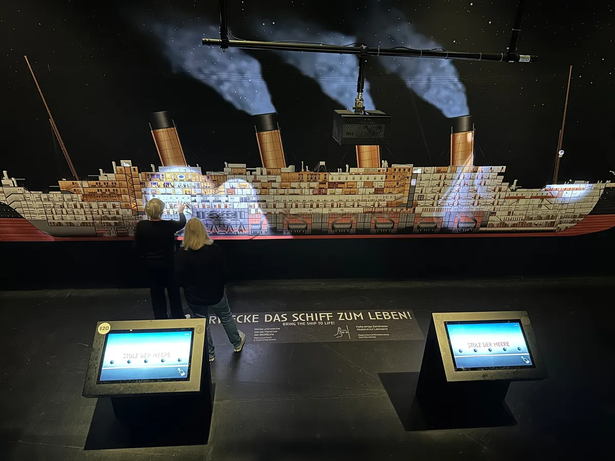 Zwei Personen betrachten ein illuminiertes Ausstellungsstück der Titanic mit interaktiven Bildschirmen.